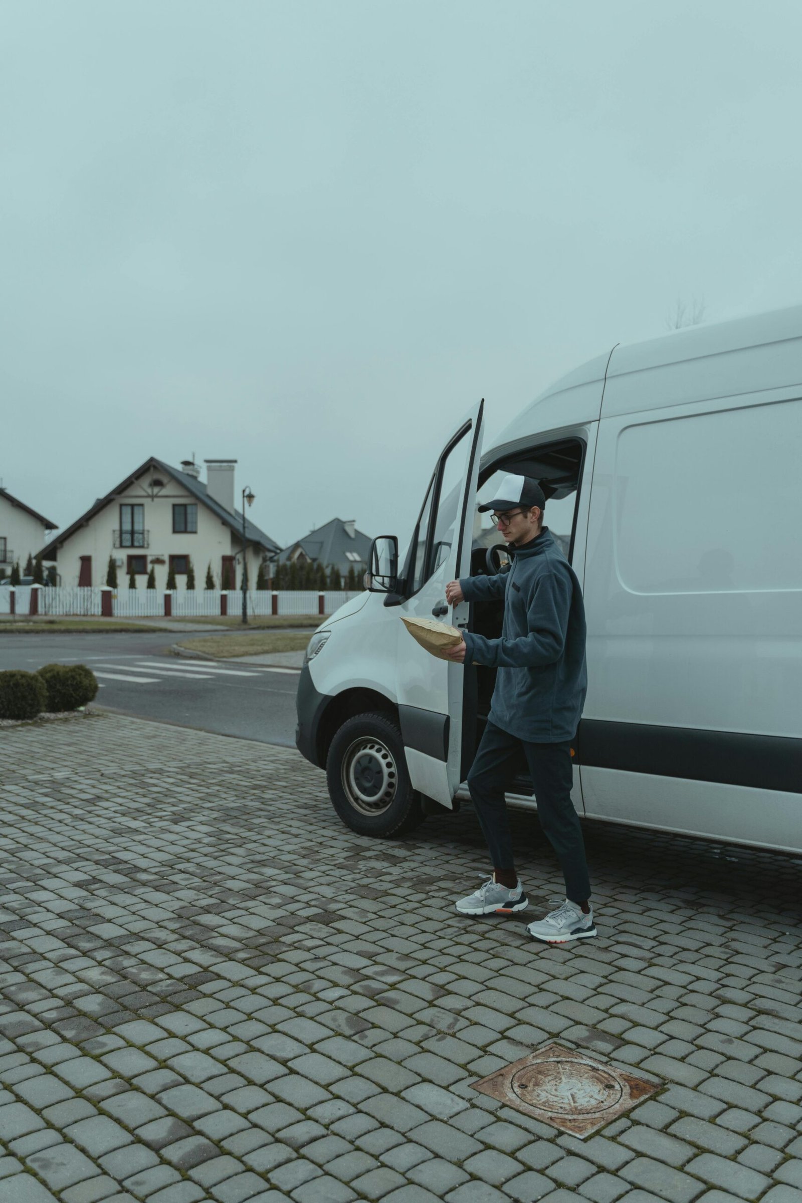 A delivery worker in casual attire carrying a parcel from a white van on a suburban street.